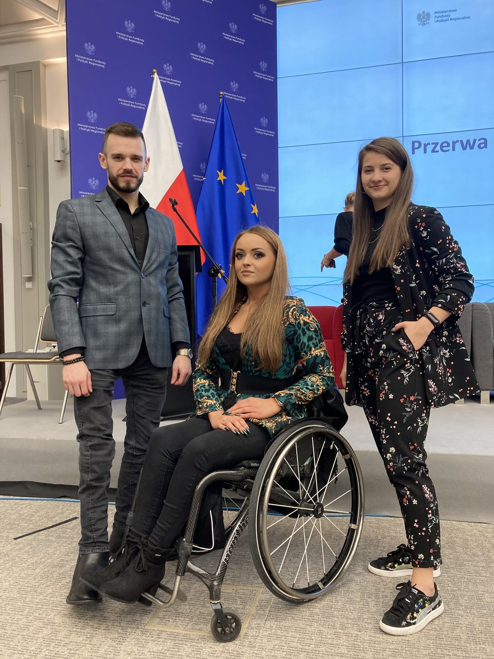 Pracownicy biura Aleksandra Zagórska, Jakub Bułacz i Monika Twardawski-Karbowiak stojący na tle podium oraz flagi polskiej i europejskiej.